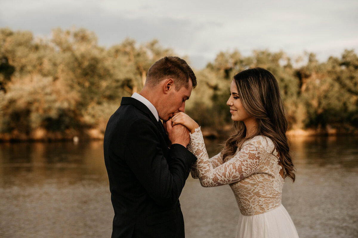 groom kissing bride's hands by a river