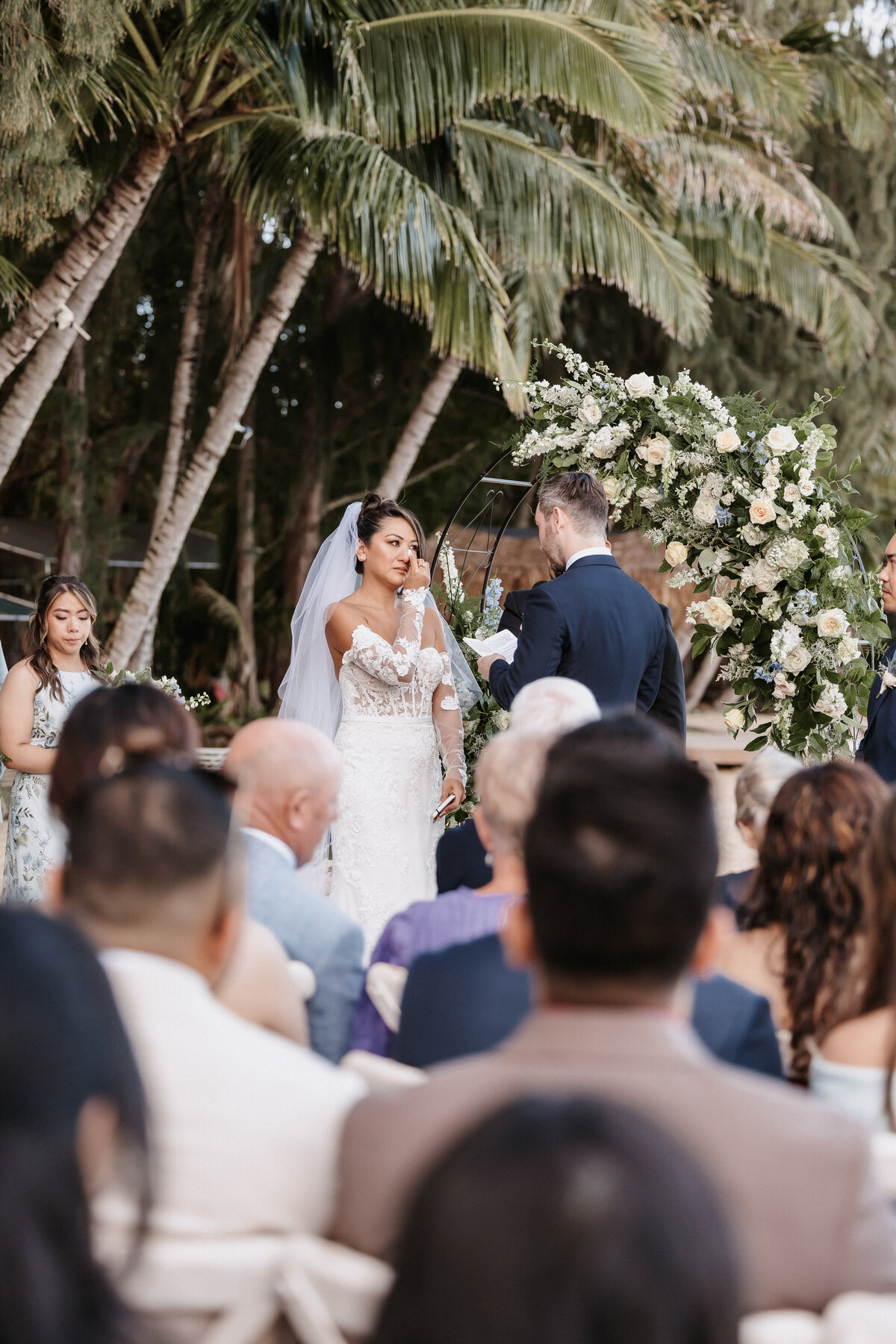 Ceremony on the beach with floral arch behind couple, bride is wiping away a tear as the groom holds the microphone reading vows