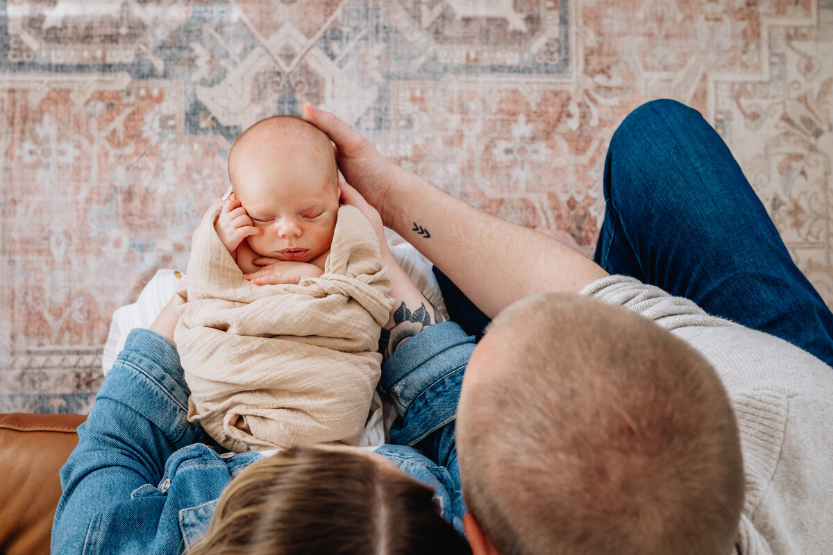A view from above of parents gazing lovingly at their swaddled newborn lying between them on a patterned rug, creating a serene family moment.