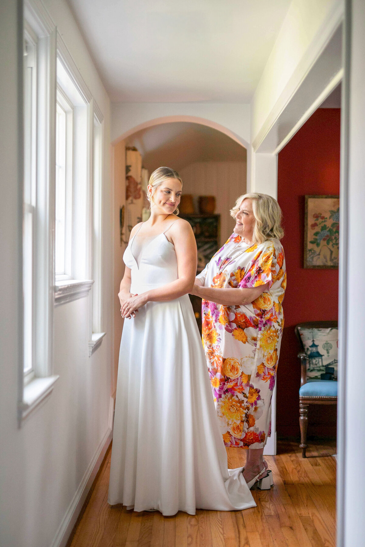A bride in a white gown stands in a sunlit hallway, smiling at an older woman dressed in a colorful floral robe who is adjusting the brides dress. They share a joyful moment together before the wedding.