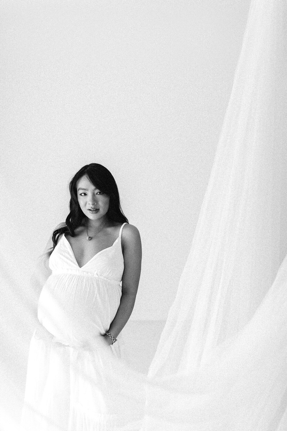 Asian woman who is pregnant stands next to white netting, wearing a white dress and looks at the camera
