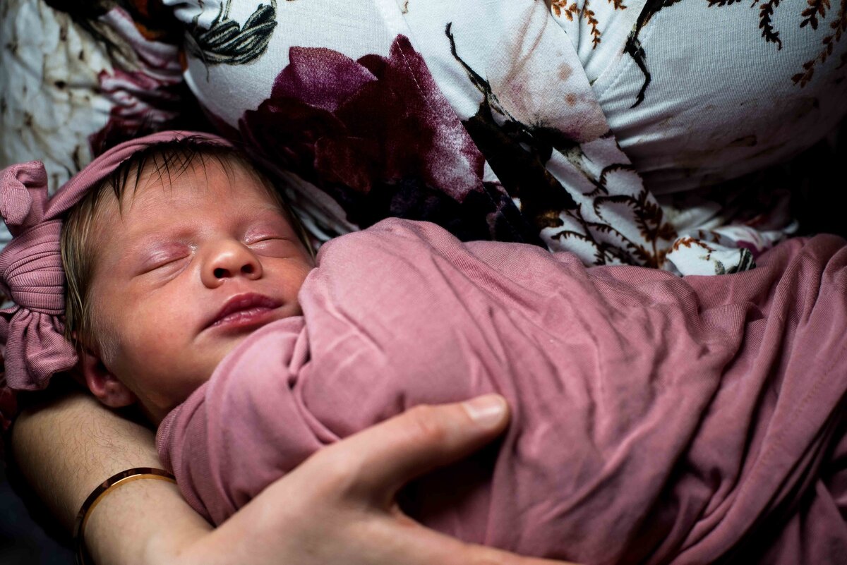 a sleeping newborn is swaddled in a pink blanket being held by her mother