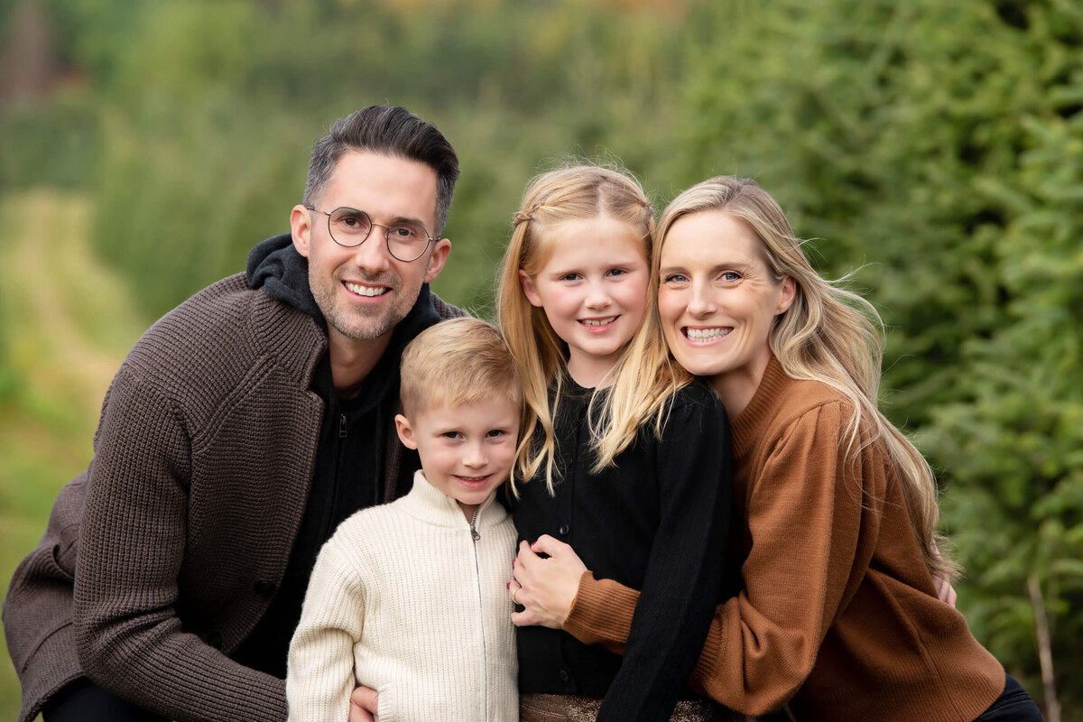 Mom and dad crouched down hugging their son and daughter in the Christmas tree farm for their Burlington family photographer session