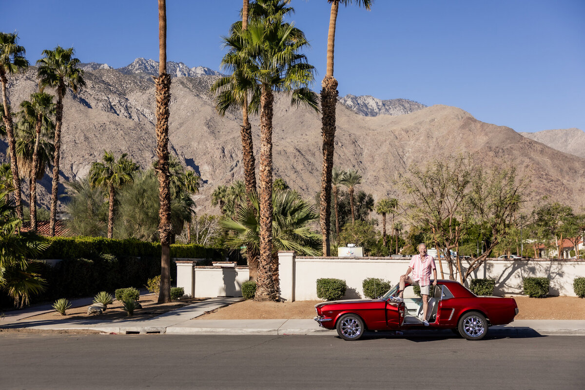 Individual posing confidently beside a red vintage convertible with palm trees and desert mountains in the background, captured in Palm Springs.