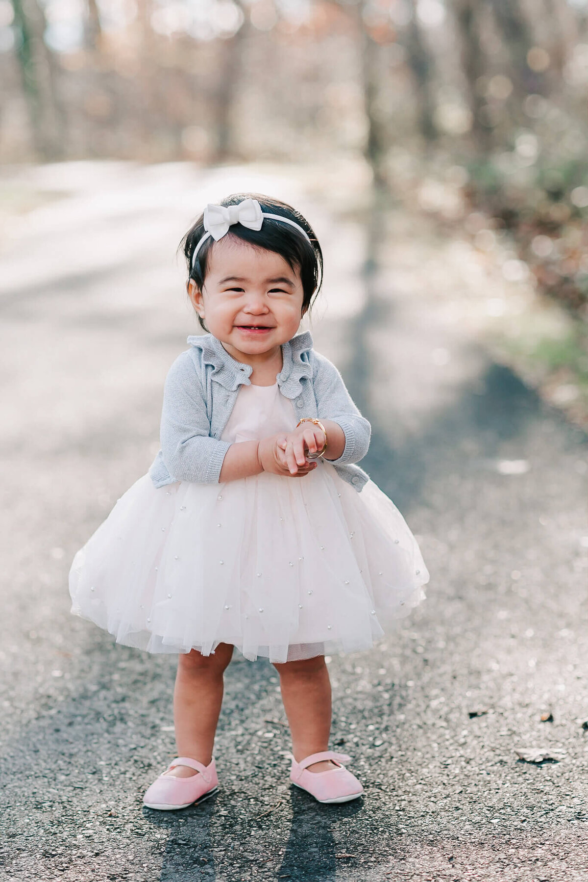 Sweetly smiling one year old girl wearing a pale pink tulle dress and grey cardigan