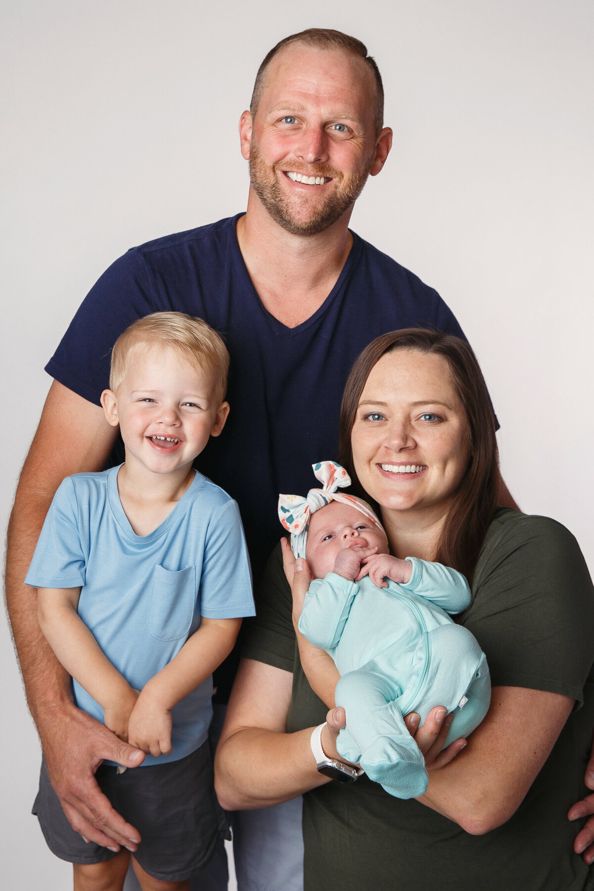 Family of four posing on a white background with mom, dad, 3 year old boy and newborn girl