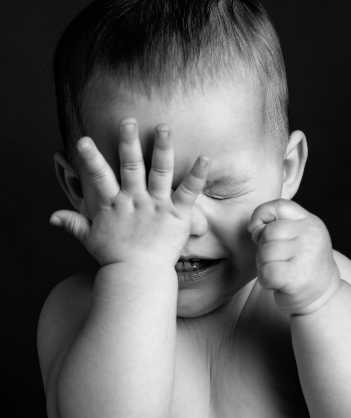 A baby playfully covers his eyes with his hands, showing a cute expression of surprise or shyness.