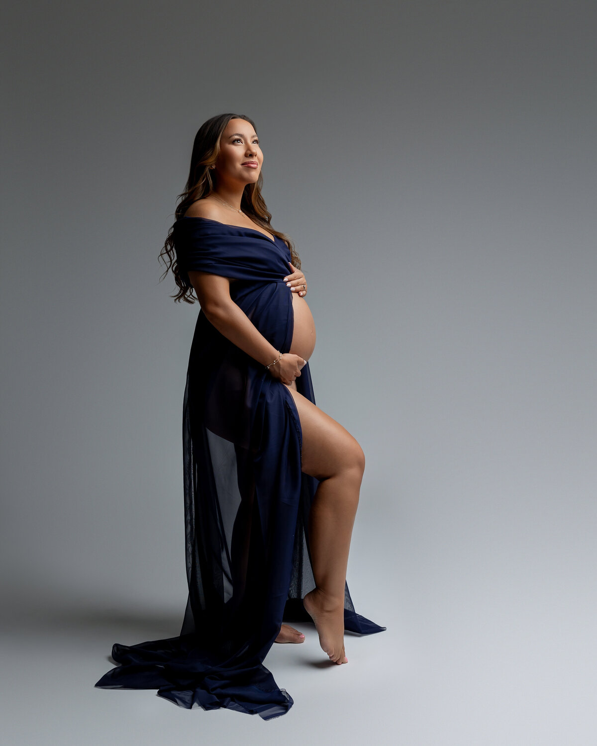 Striking side profile of a glowing expectant mother in a sheer navy gown, confidently showcasing her baby bump against a soft, modern backdrop. Captured by Inland Empire maternity photographer Kaitlyn Dawn Photography.
