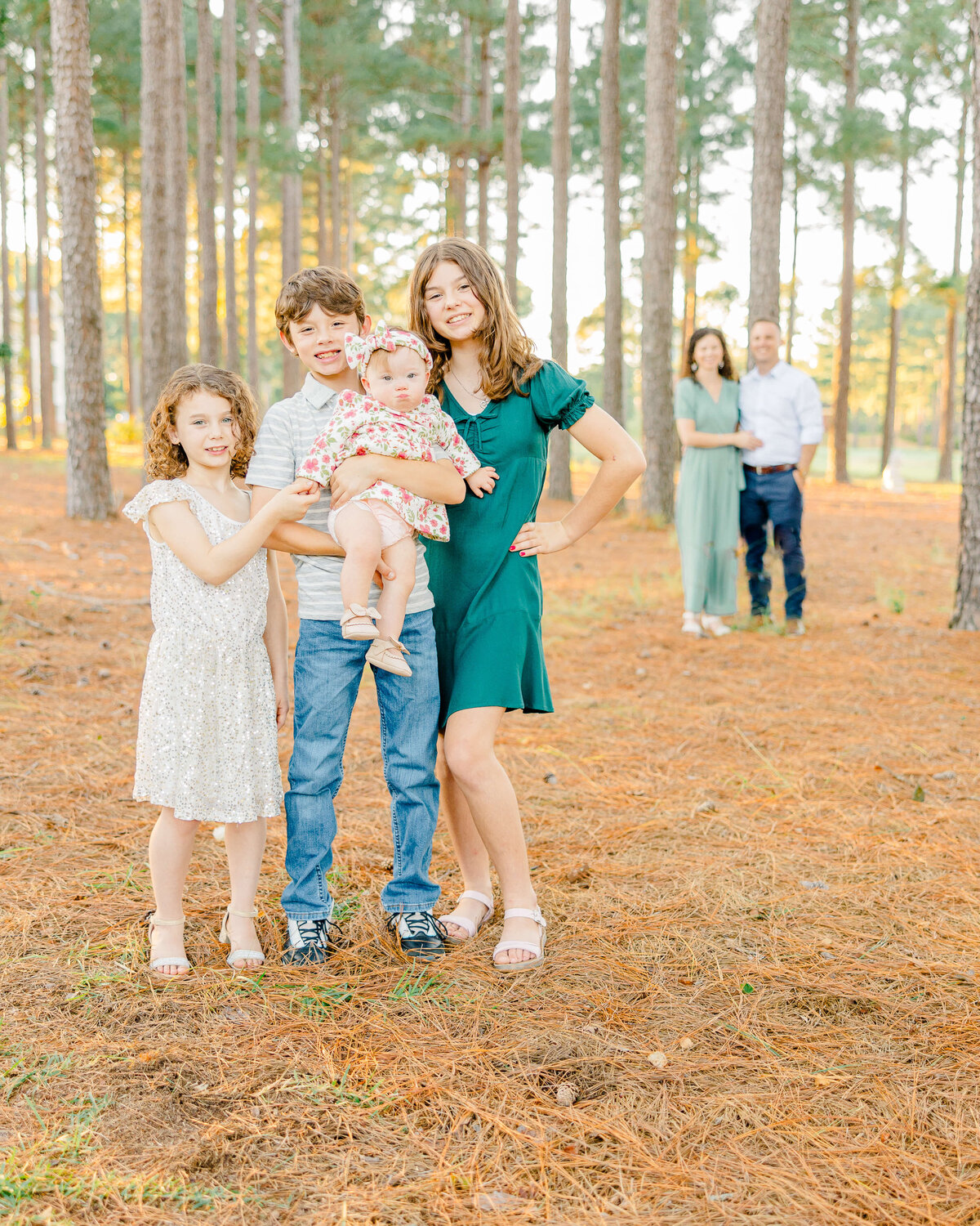 In the serene pine forest, captured by a Fayetteville NC family photographer, an older girl in a green dress strikes a pose with her hand on her hip. Nearby, a boy in a striped shirt holds a baby while a young girl in white stands beside them, and two adults linger softly in the background.