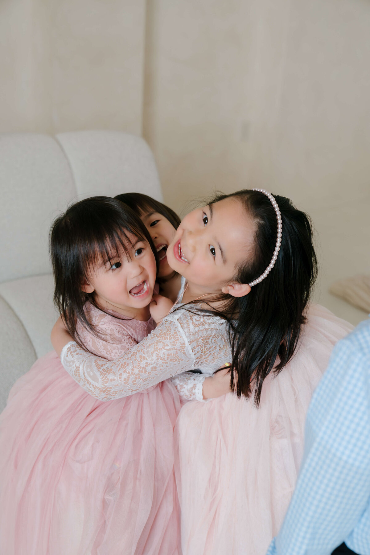 Three sisters dressed in pastel dresses share a happy hug on a modern cream couch.