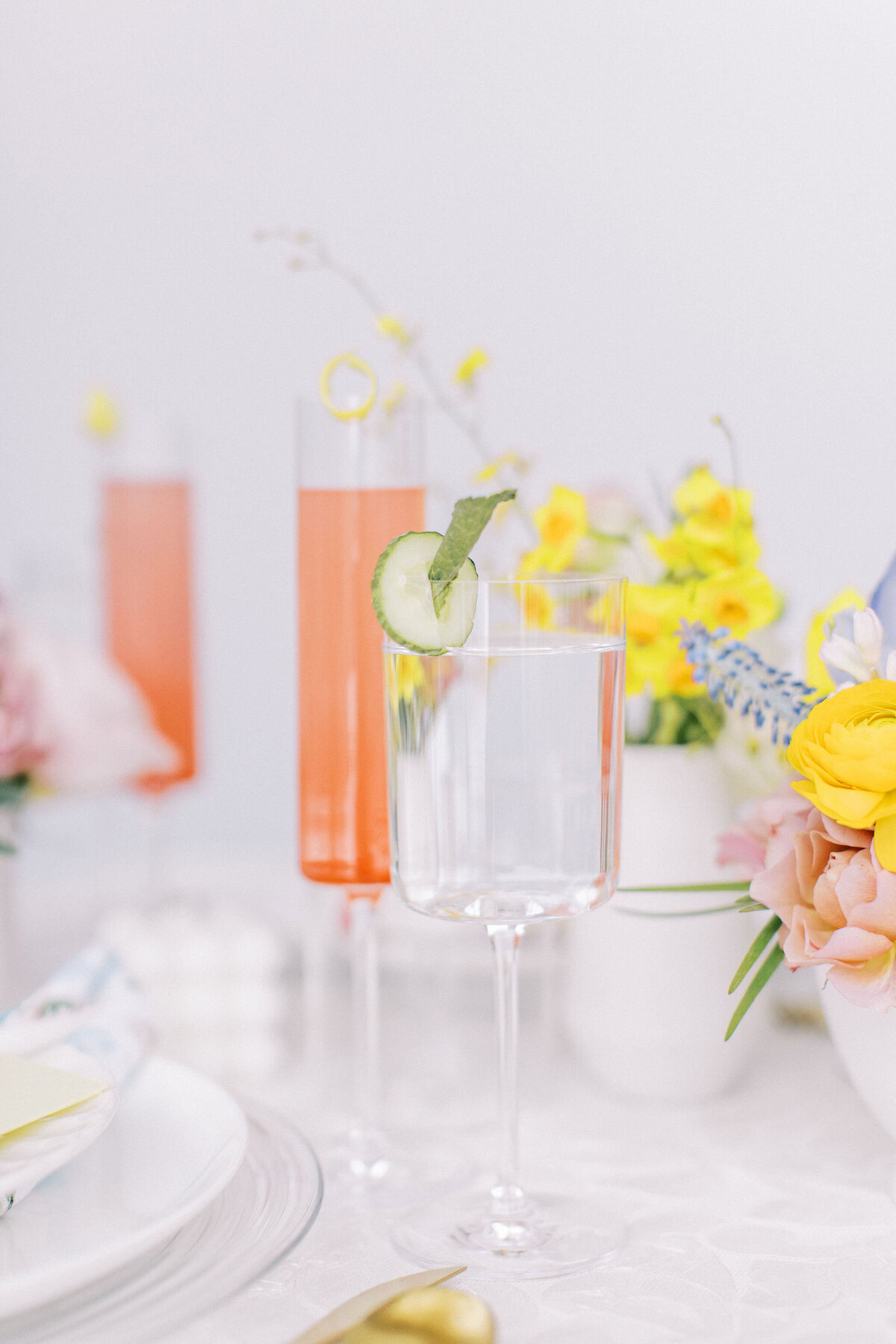 A table is set with a clear glass of water garnished with a cucumber slice and mint, alongside two pink beverages in tall glasses. Bright yellow flowers are in the background, creating a serene atmosphere perfect for weddings in Banff Alberta.