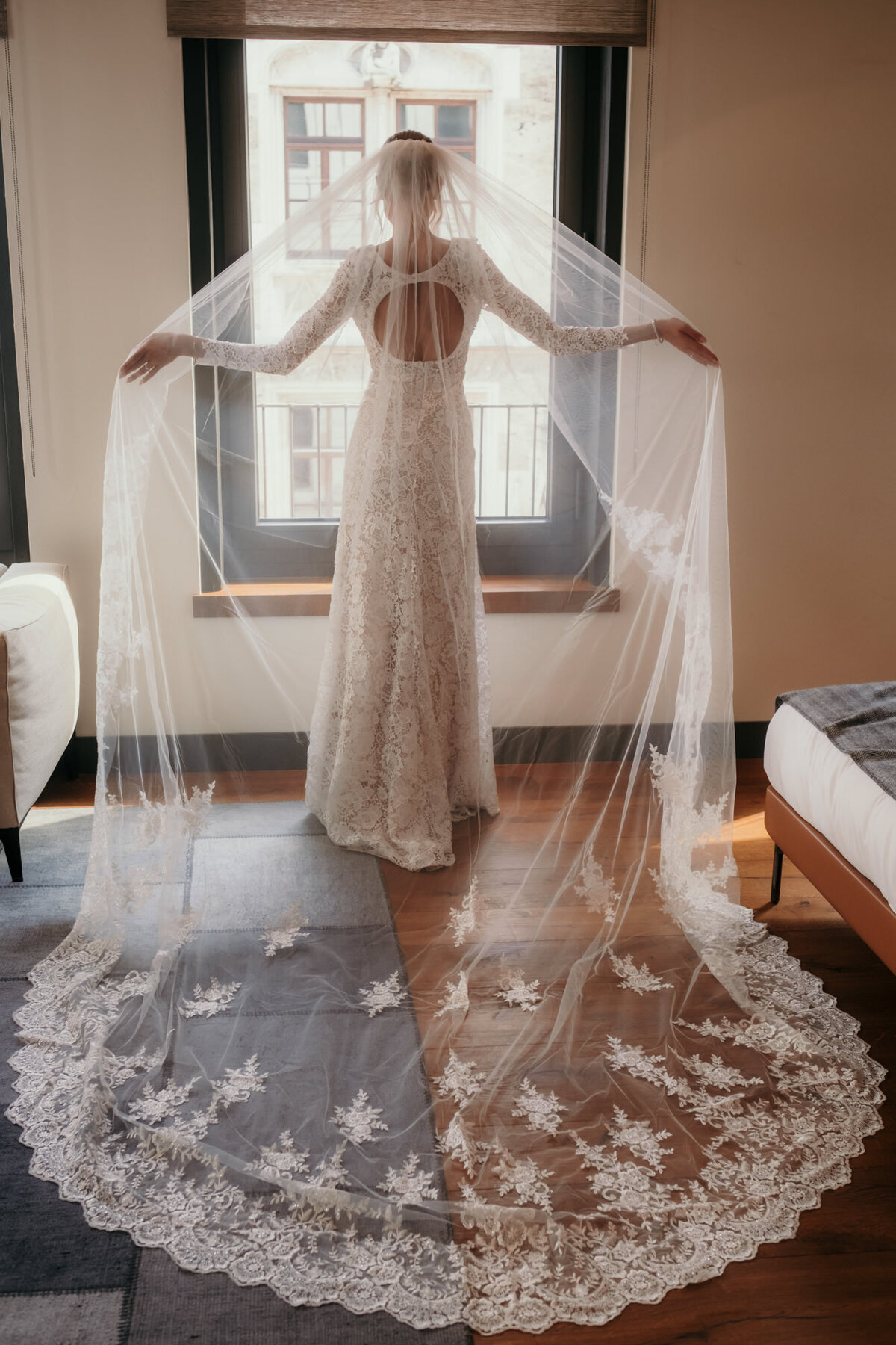 The bride in her lace-covered dress stands in front of a window holding up her big gourgeous veil.