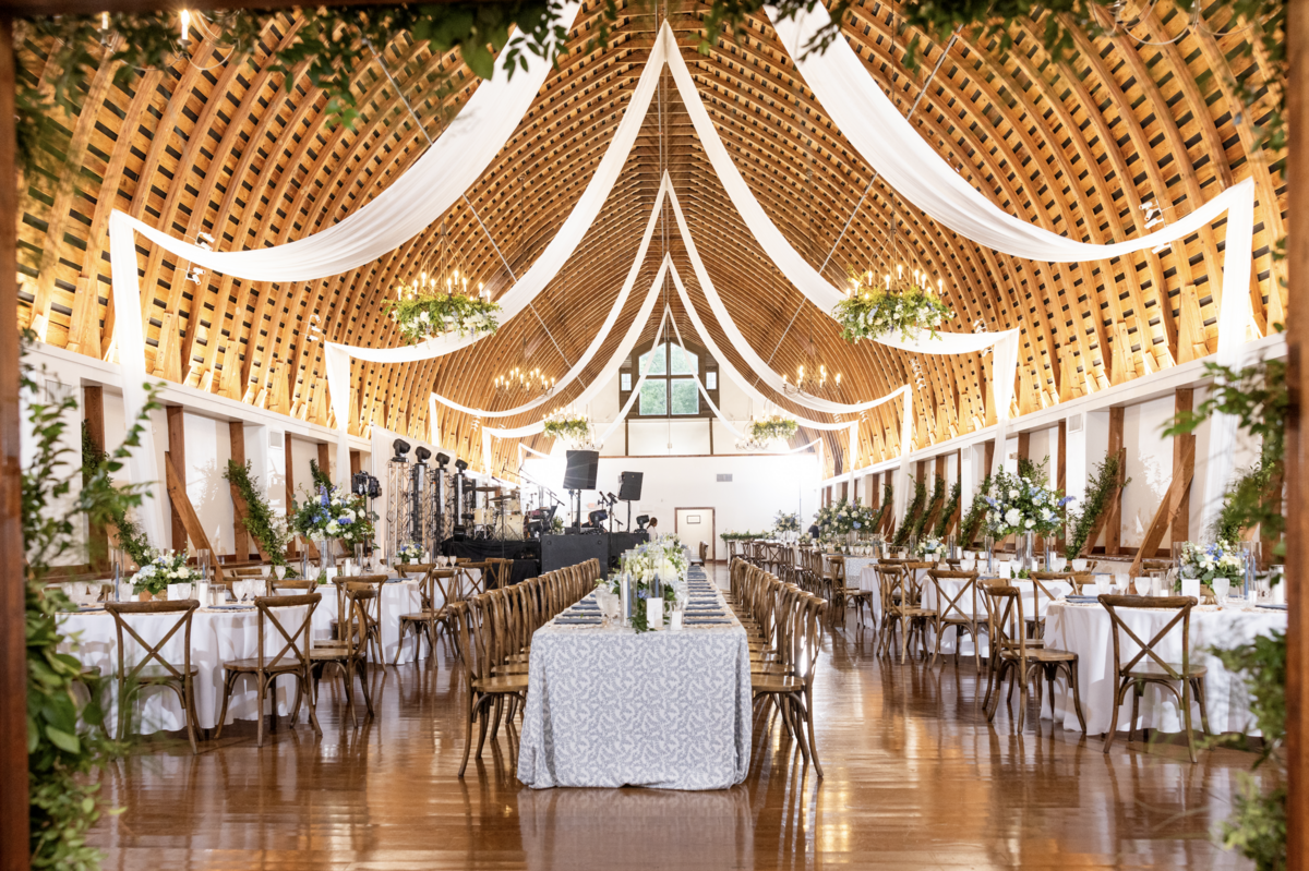 Spring wedding reception at WinMock at Kinderton, designed by Charming Carolina — wide view of the Loft with fairy lights, white ceiling draping, greenery-covered columns, and chandeliers adorned with dusty blue blooms and heavy greenery.