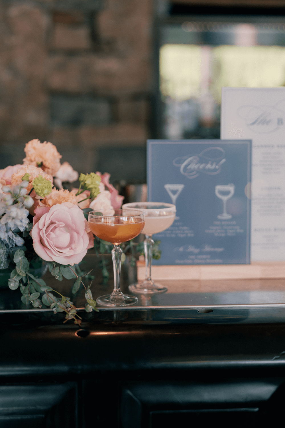 Signature cocktails displayed with a custom acrylic bar menu and pastel floral arrangement at a summer wedding at Moonlight Basin.