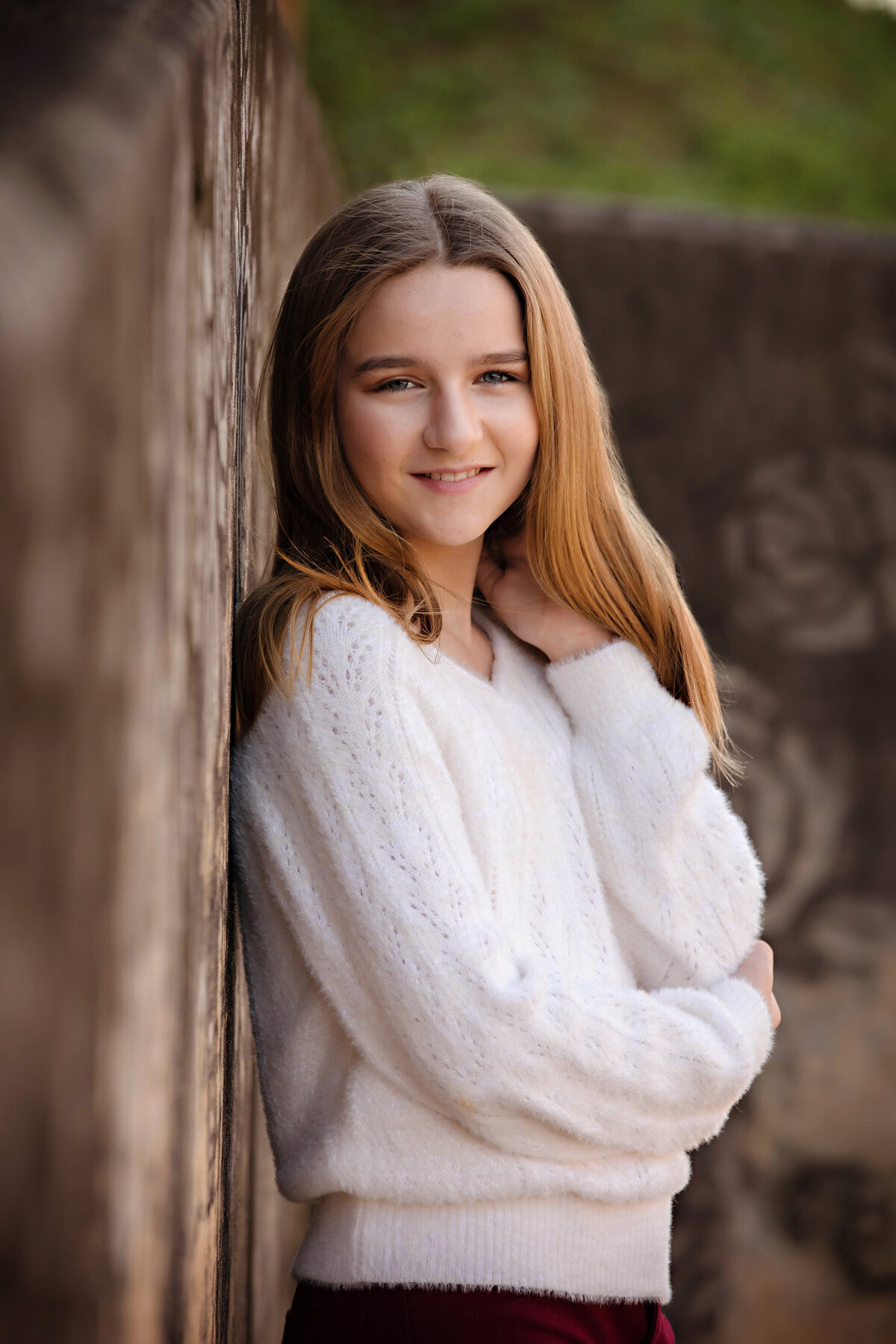 Lovely HS freshman in cream sweater and cranberry pants standing against weathered concrete wall with faint rose imprints in autumn light at Camp North End Charlotte NC