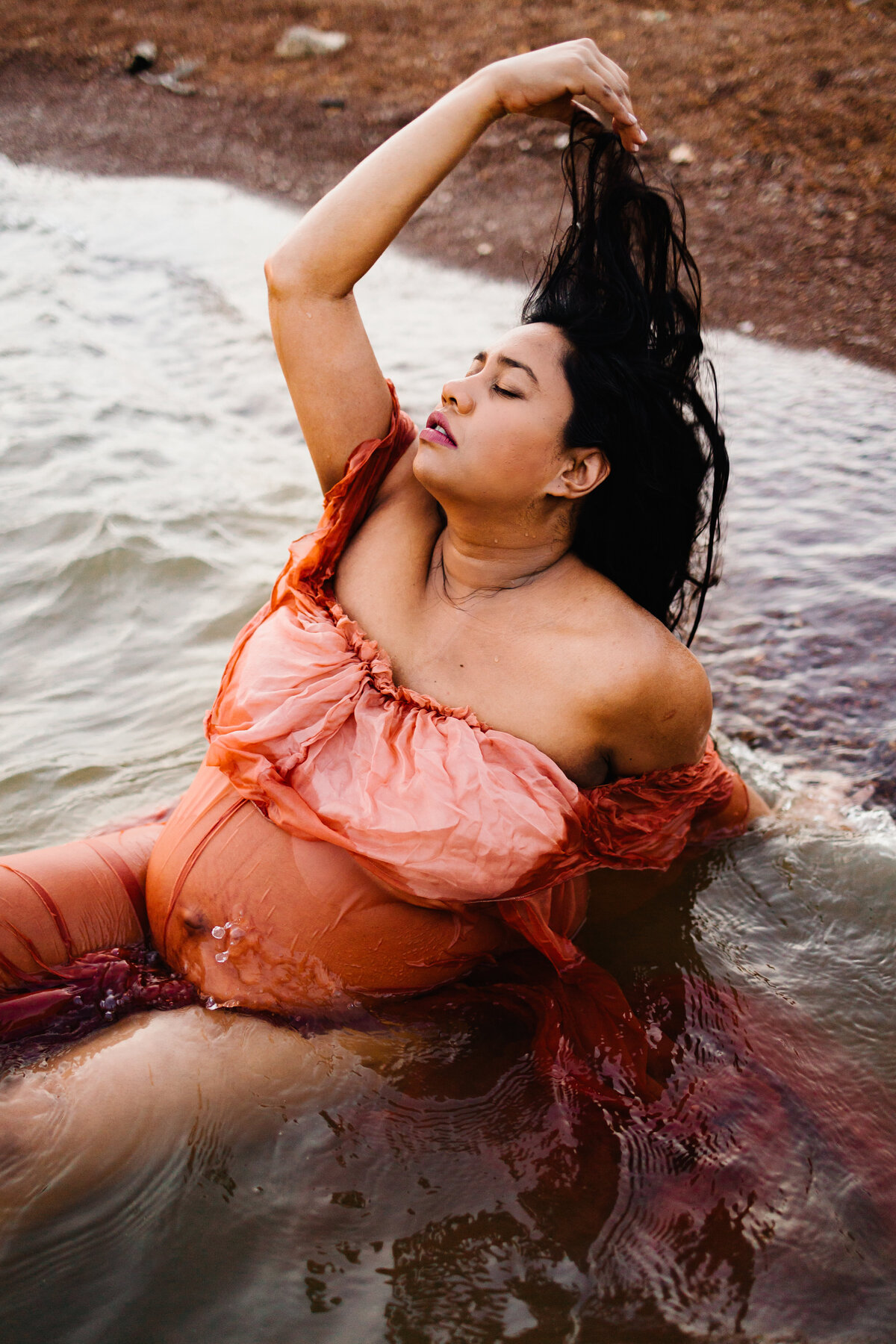 A stunning image by a maternity photographer in Albuquerque. The pregnant woman stands gracefully in a clean lake, wearing a long orange dress. She holds her hair with one hand on top, her eyes closed, capturing a serene and peaceful moment.