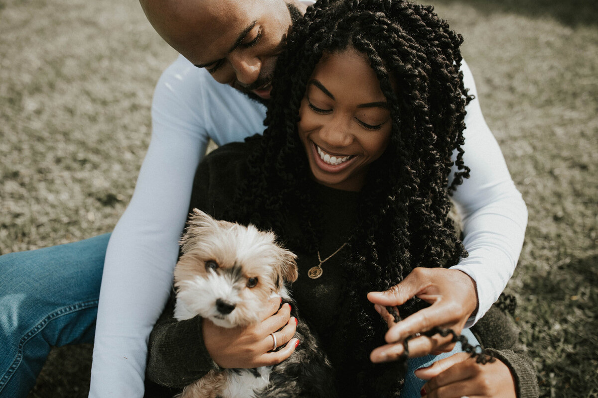 Couple sitting on the grass, the man hugging the woman from behind as she holds a small dog.