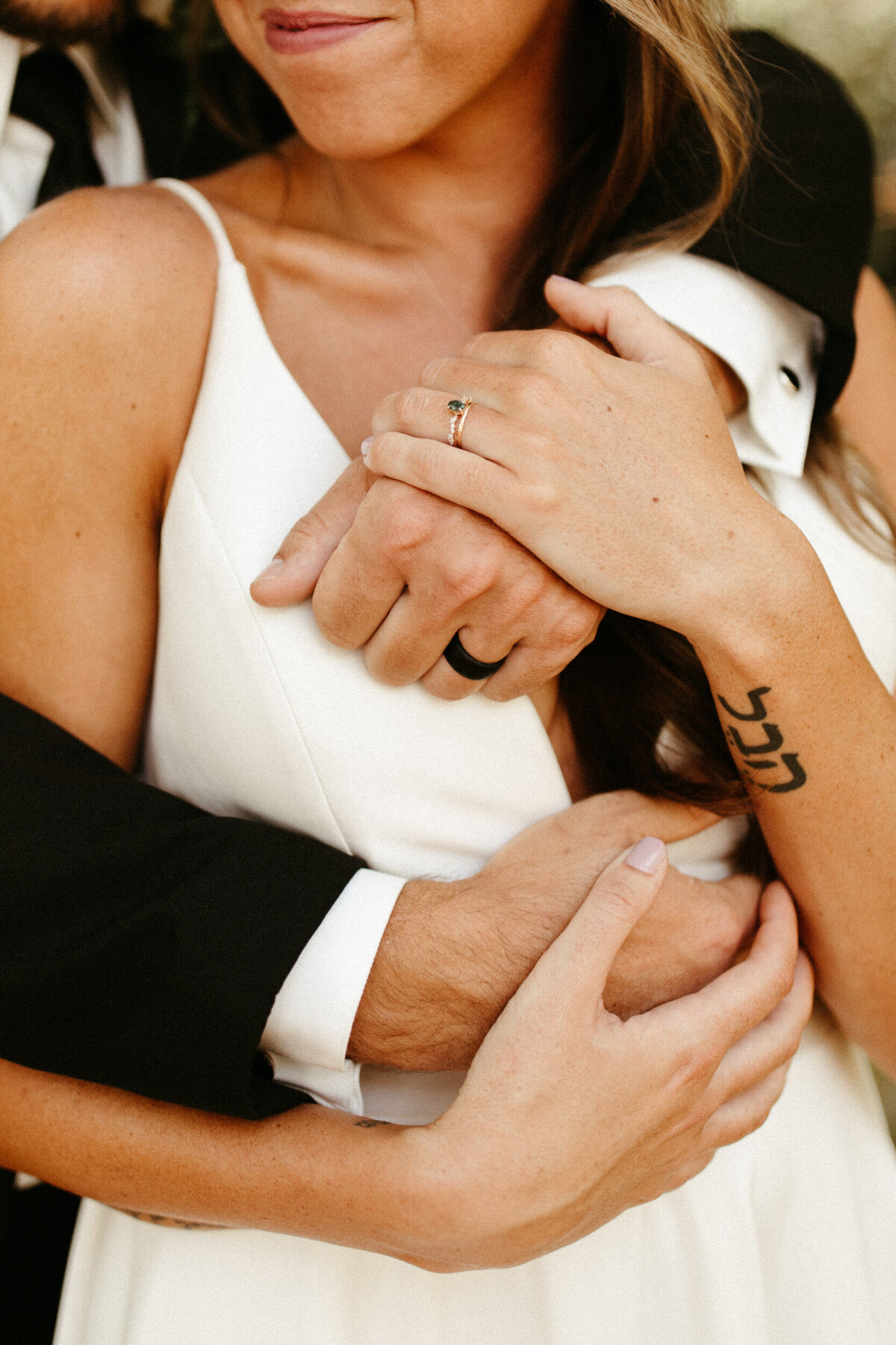 Close up of bride with emerald engagement ring holding her groom's hands as he wraps his arms around her