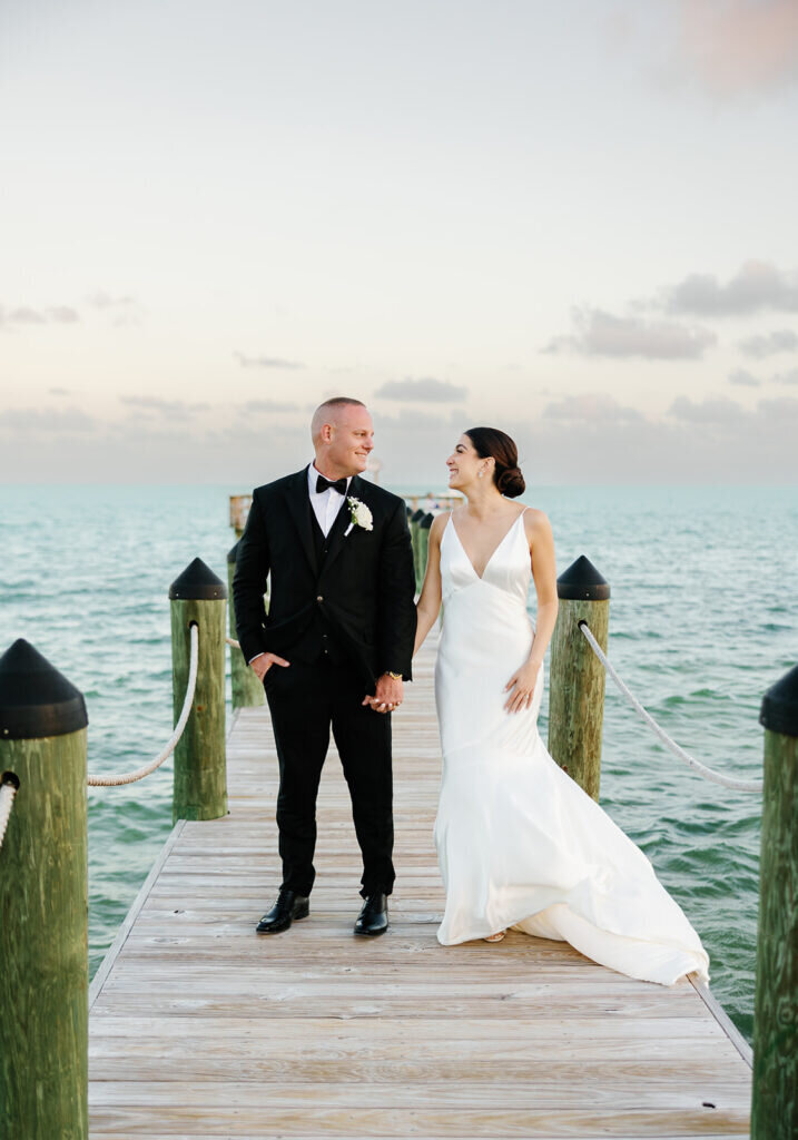 A romantic portrait of the newlyweds strolling along a wooden dock, with the ocean stretching out behind them under a pastel evening sky. Captured by Claudia Amalia Photography.