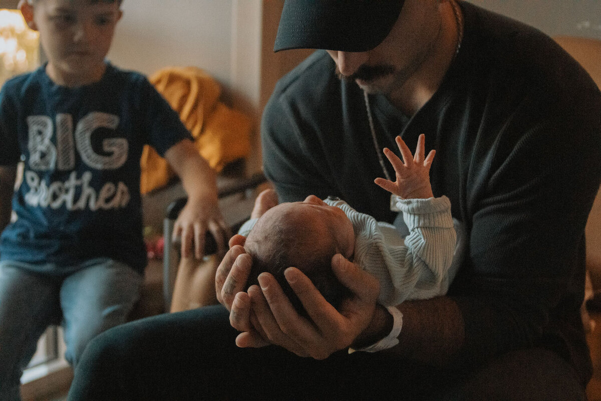 Father holds his newborn close while a toddler looks on in the background