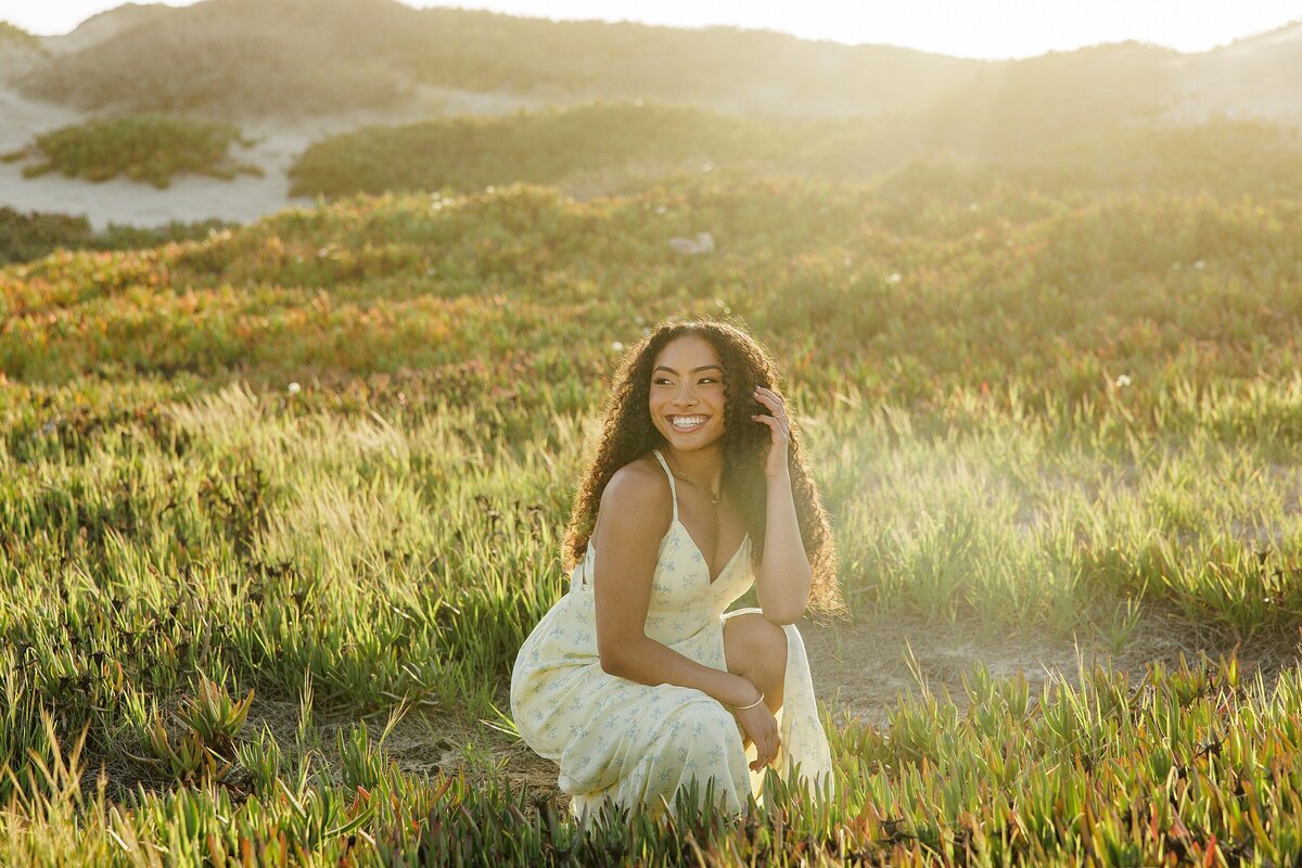 Editorial-style senior portrait of a girl in a yellow dress, sitting in golden-hour light among wild grasses and greenery near the Southern California coast.