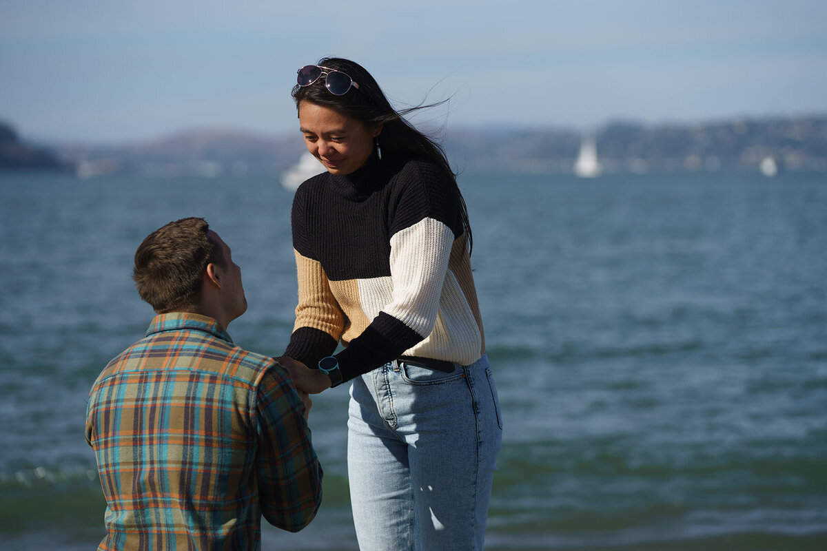 Man on one knee proposing to a surprised woman at the beach with sailboats in the background
