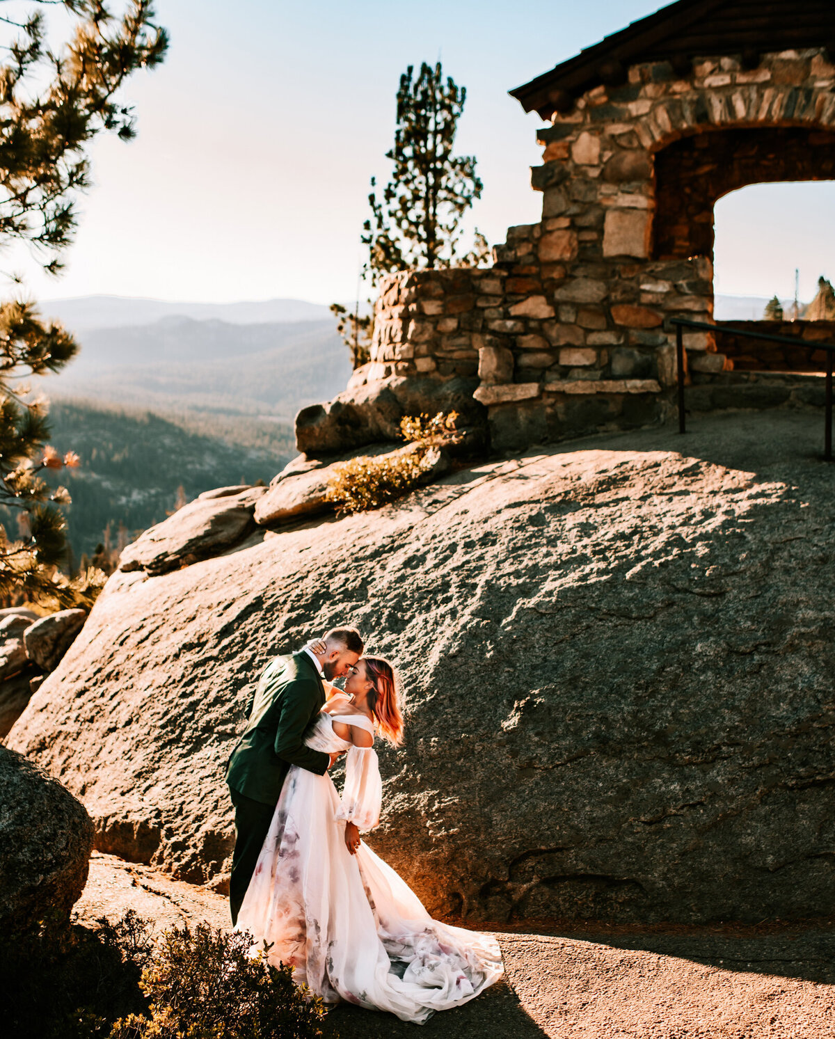 Couples Photography, Man in a blue blazer kissing and holding woman in a wedding dress at the bottom of a large rock