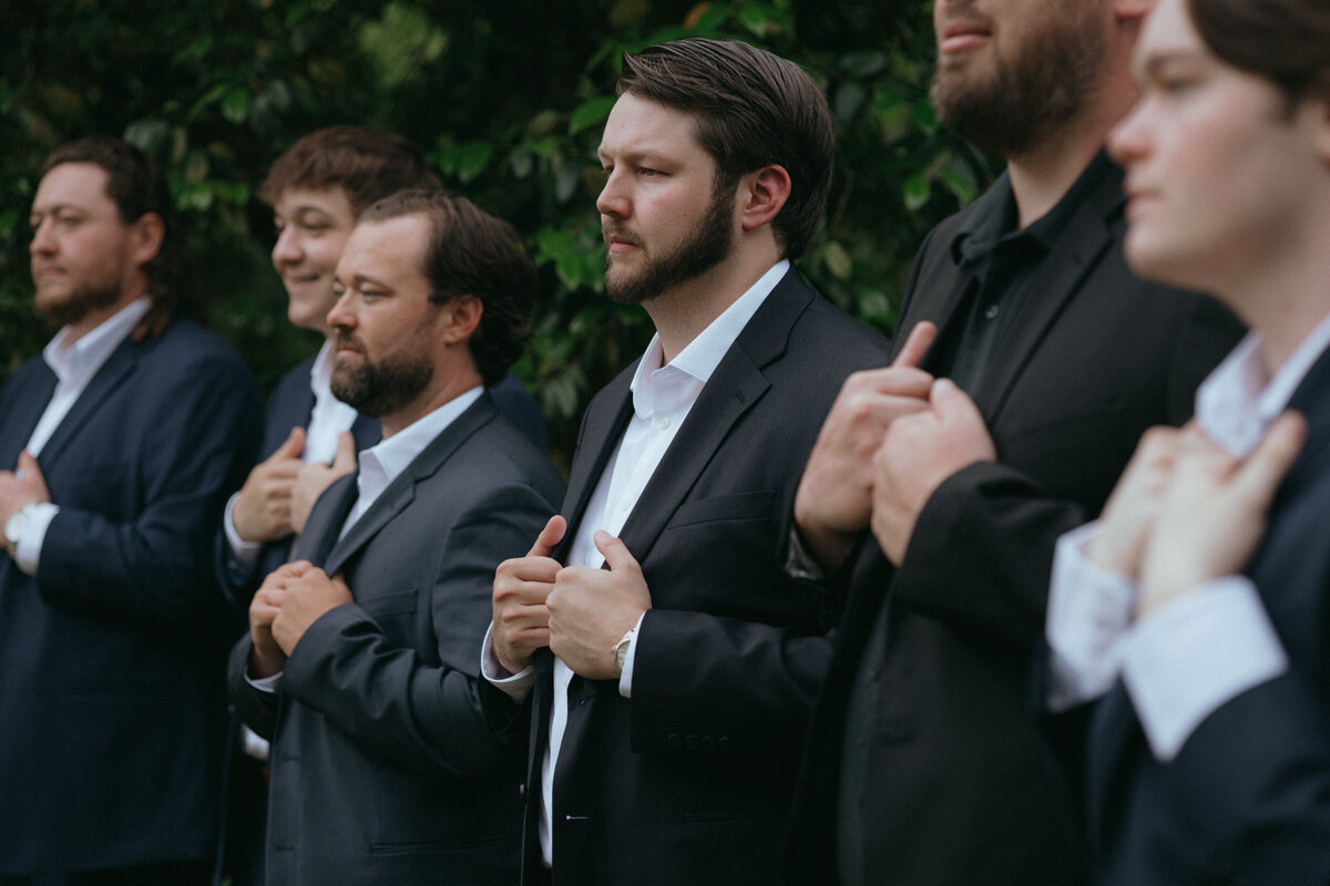 Groomsmen line up for a photo holding their navy blue suit jackets. They have a serious look on their face and the focus is on the groom. Surrounded by greenery at Seibels House and Gardens in South Carolina..