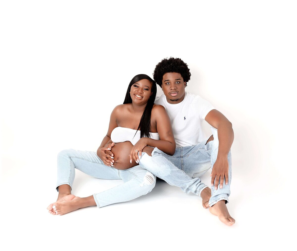 A couple sitting on white backdrop wearing jeans and white top for their maternity session.