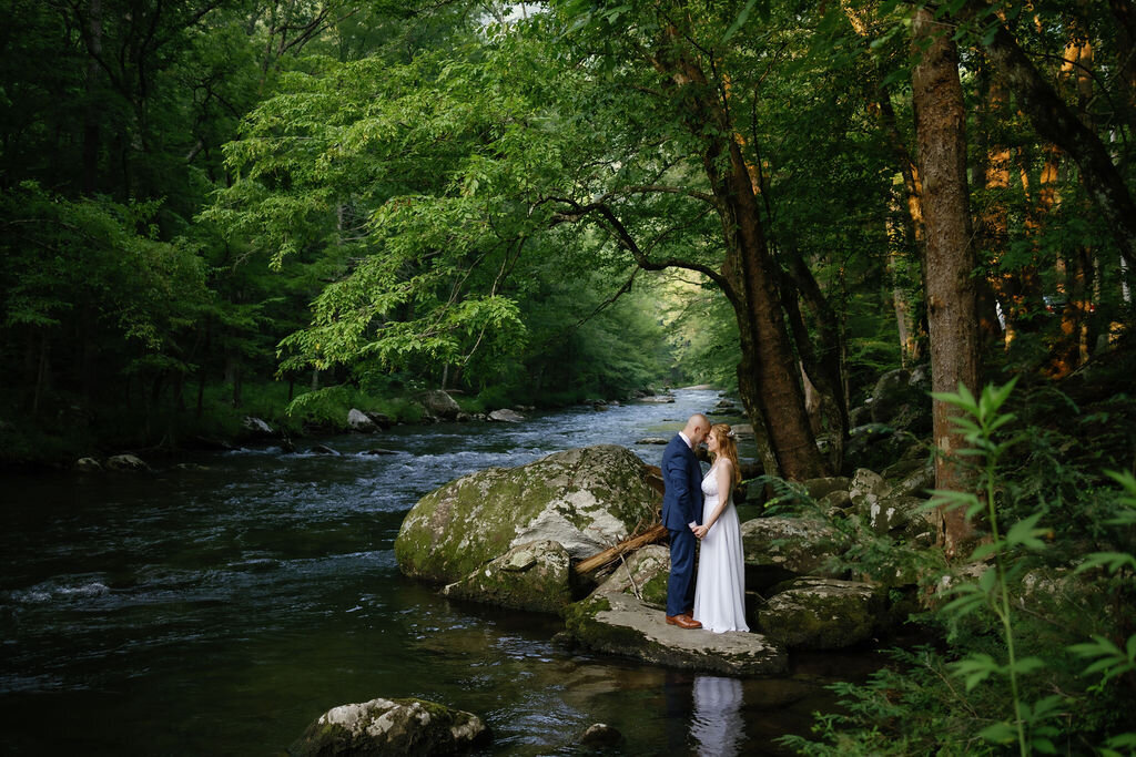 Gatlinburg elopement planner has a forrest wedding with bride and groom holding hands and leaning into each other while standing on the bank of a river
