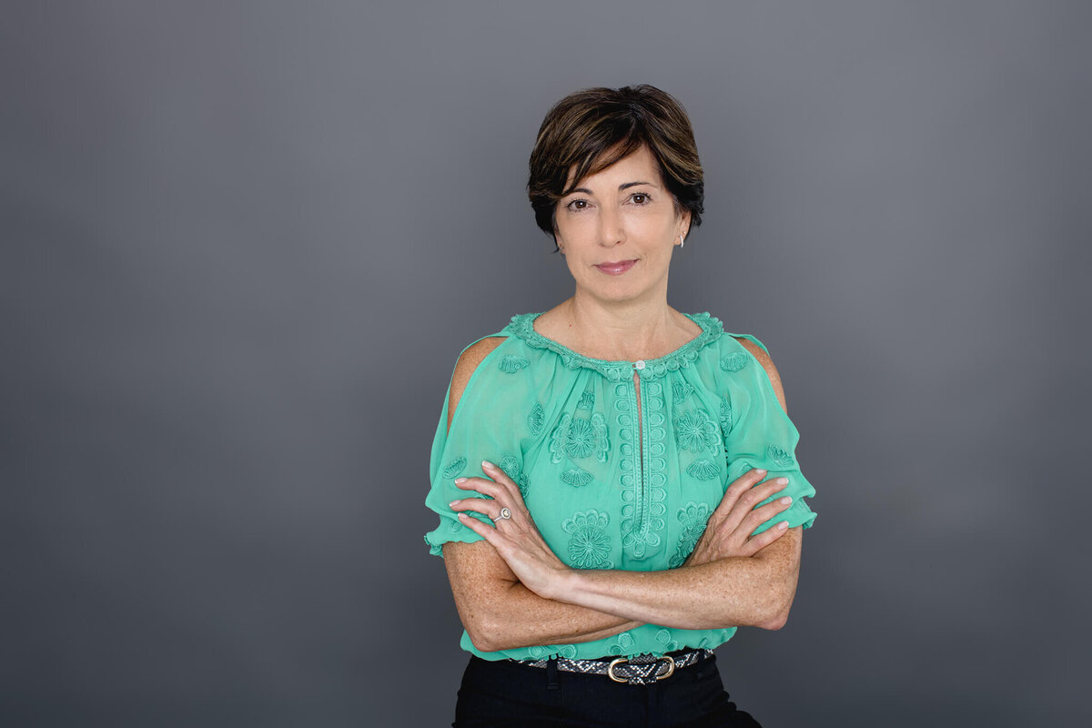 A woman with short black hair wearing a light green blouse sitting in front of a dark grey backdrop with her arms crossed over her chest, smiling with only her lips at the camera.