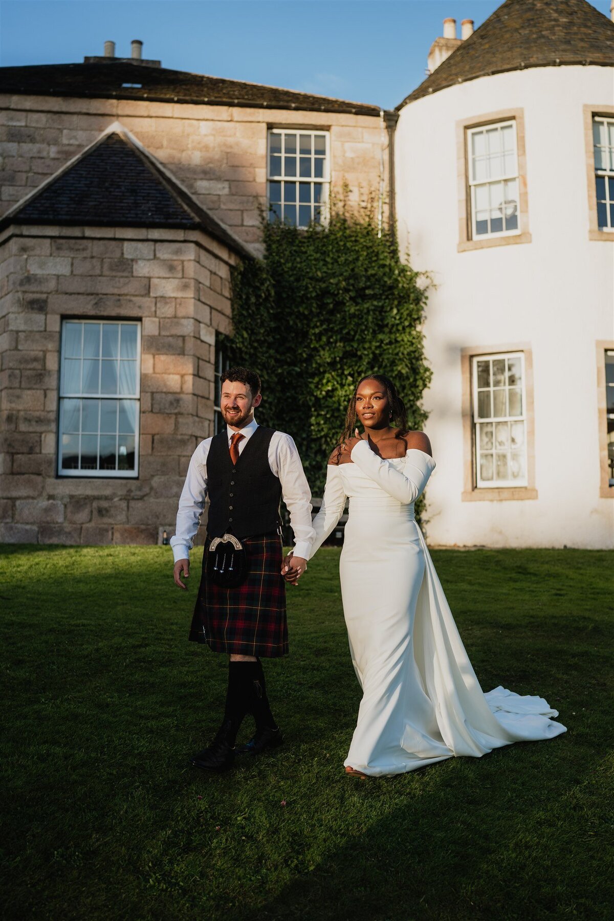A bride and groom hold hands and walk down a gentle grass hill. White cladding can be seen on the walls behind them. They look off to the distance as the sun begins to set. A golden light is seen in the photograph.