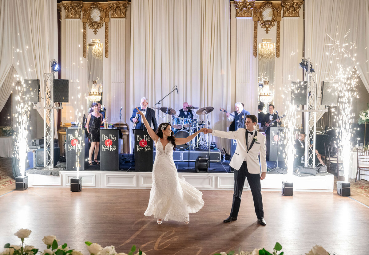 A bride and groom perform their first dance in an elegant ballroom. The bride wears a white gown, and the groom is in a white tuxedo. A live band plays in the background, and sparklers light up the scene.