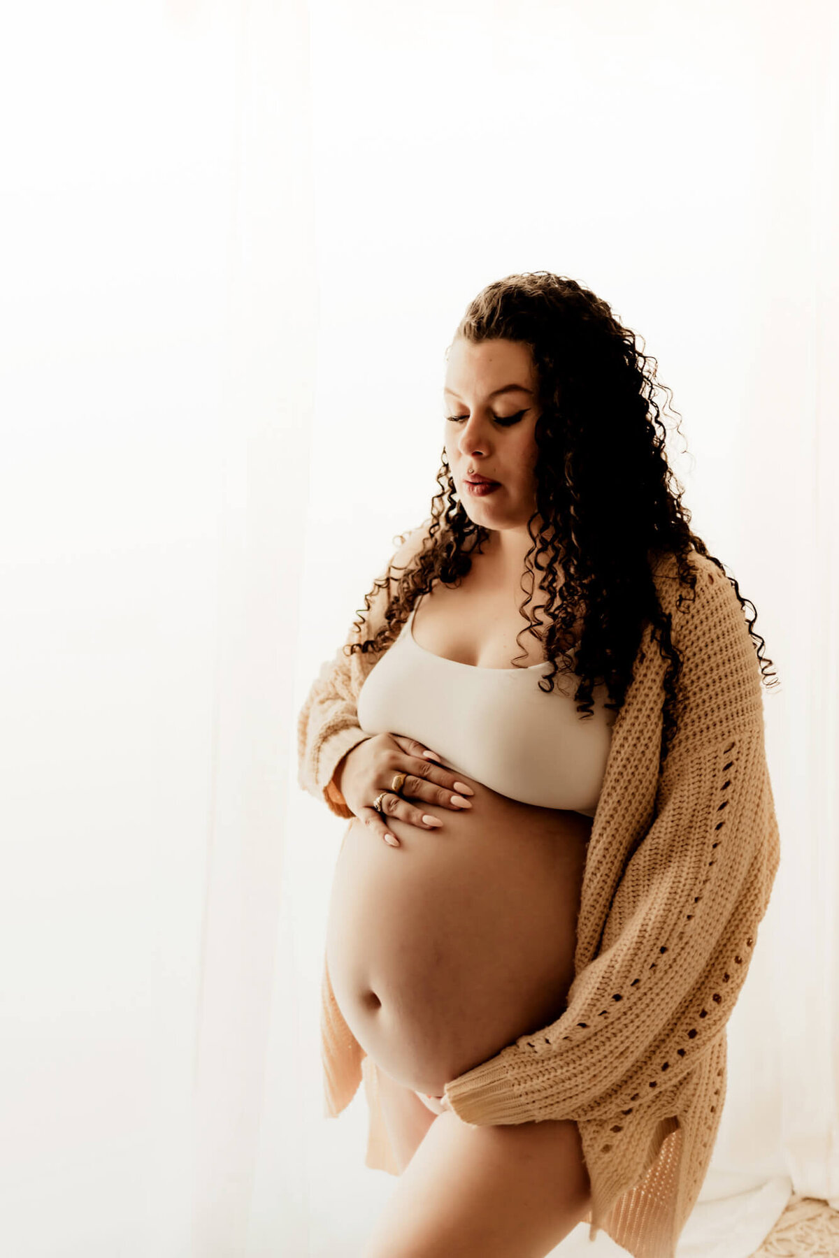 Pregnant mother wearing a white bralette and beige sweater looks at her baby bump. 