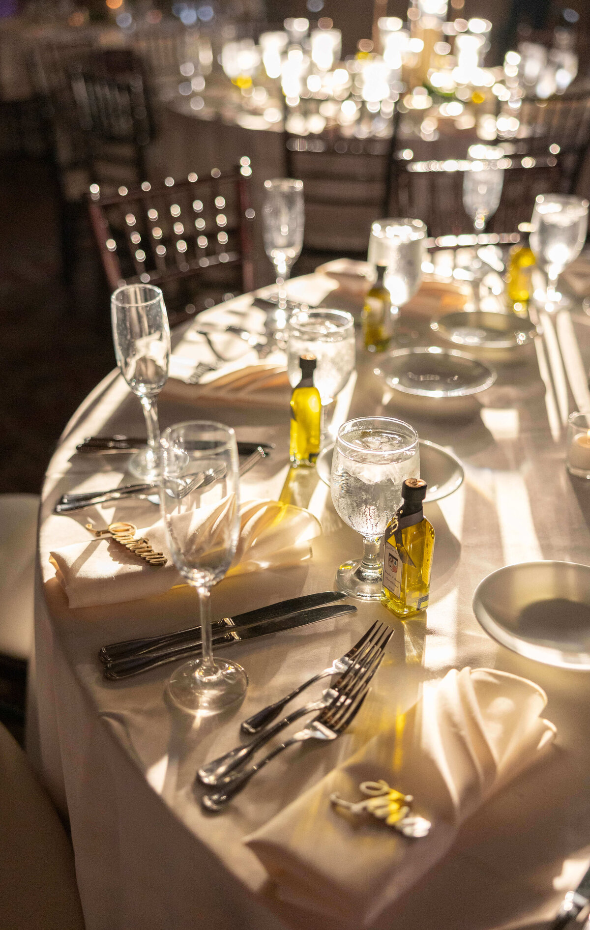 A round table is elegantly set for a formal event, featuring folded napkins, wine and water glasses, small olive oil bottles, and cutlery. Warm lighting creates a cozy atmosphere, highlighting the polished silverware and white tablecloths.