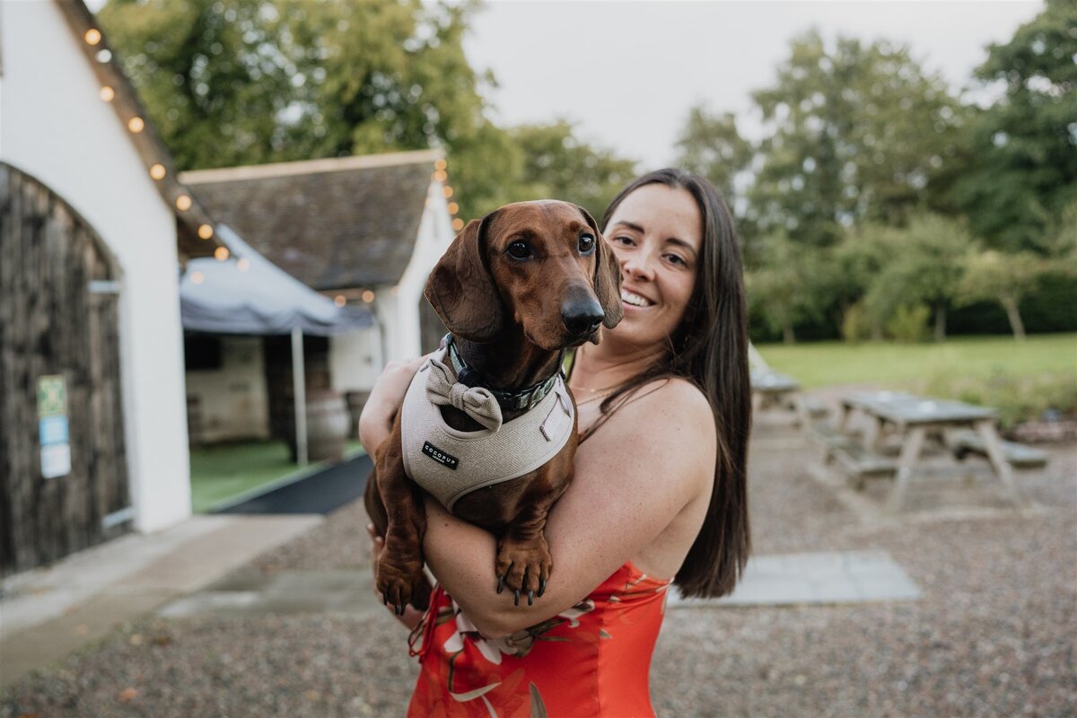 A wedding guest wearing a dress holds a sausage dog in her arms, she poses the dog for a photograph.