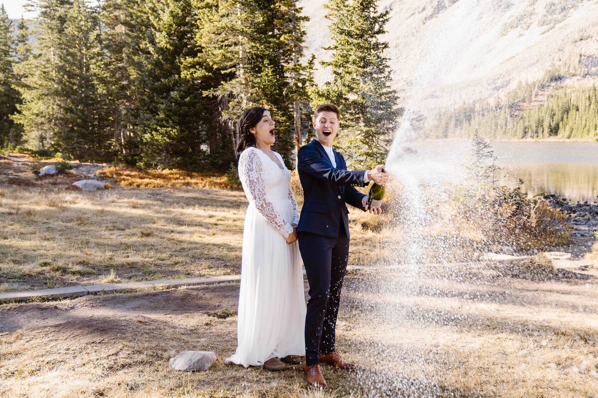Two brides pop champagne beside Mitchell Lake, celebrating with a spray against a mountain backdrop.