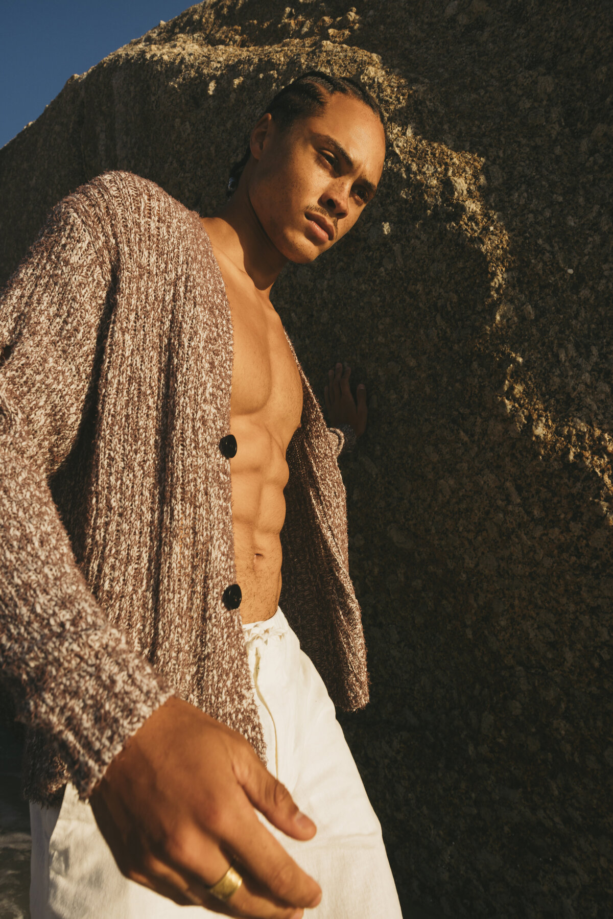 Male model Timothe standing against a large sunlit rock, wearing cream linen shorts and a loose brown knit cardigan, with defined abs visible, captured during golden hour in Cape Town.