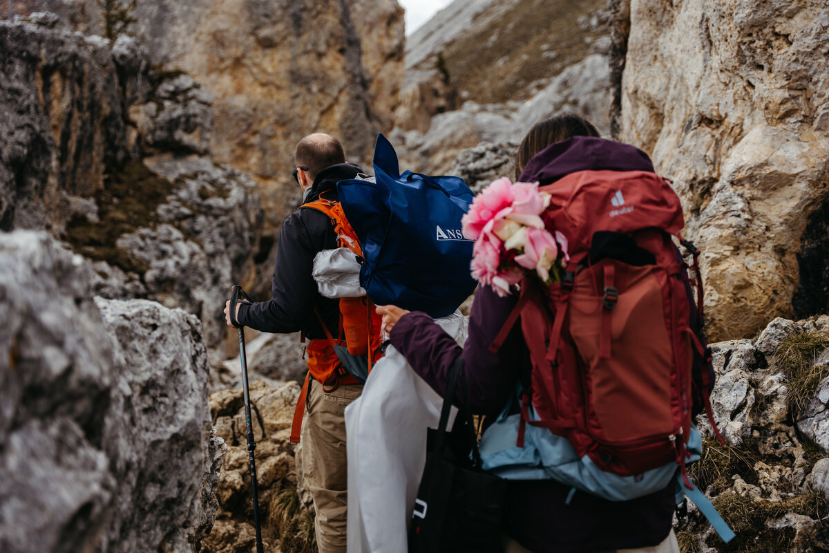 Groom helping bride climb rocky trail with backpacks
