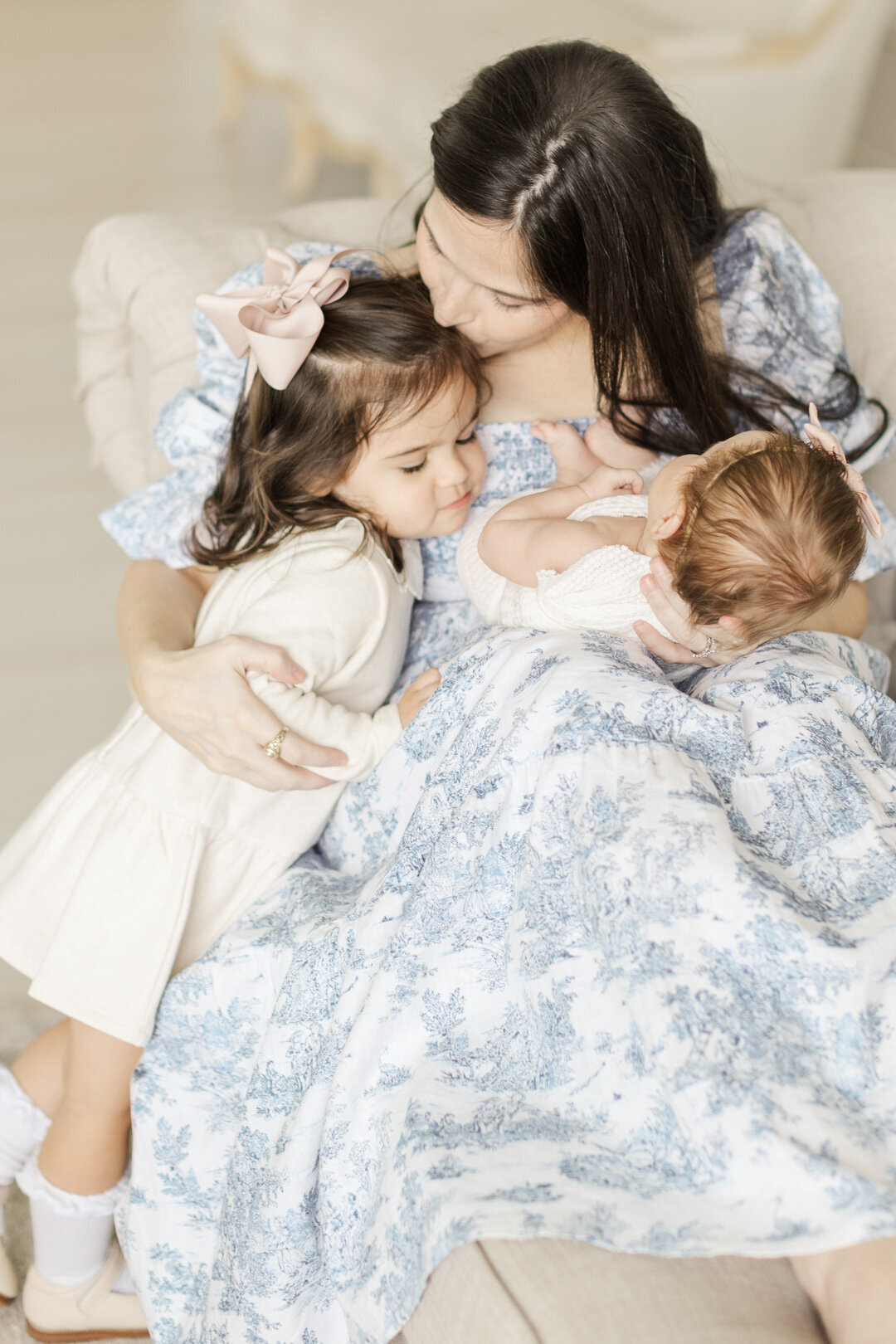 mom kisses toddler daughter on the forhead while holding baby sister on a sofa for katy newborn photo session