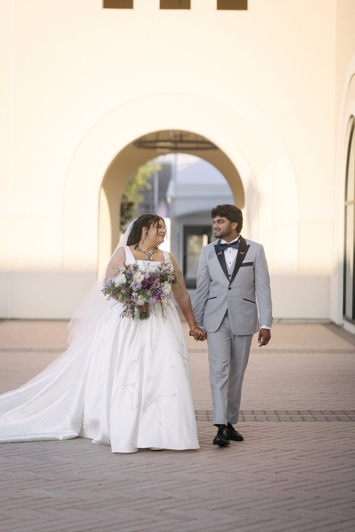 A joyful bride and groom walk hand-in-hand in elegant American wedding attire after their Indian wedding ceremony. The bride wears a flowing white gown with floral embroidery and holds a lush bouquet of purple and ivory blooms. The groom looks dapper in a light gray tuxedo with black lapels. Captured in an open-air architectural setting, this candid moment blends cultures and styles effortlessly. Photographed by Phavy Photography in a true-to-color, editorial-meets-documentary style that celebrates authenticity, diversity, and modern love.
