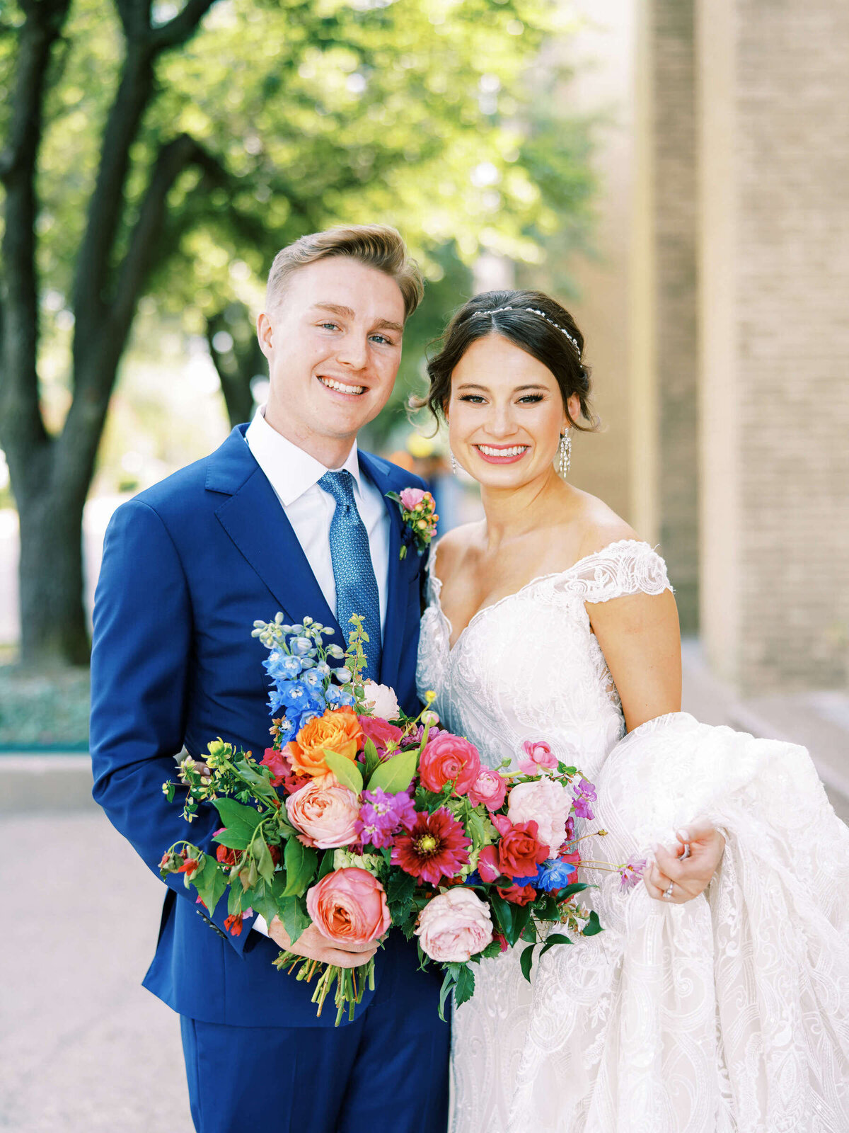 Glowing summer bride hugs handsome groom holding vibrant bouquet outside Fort Worth wedding venue