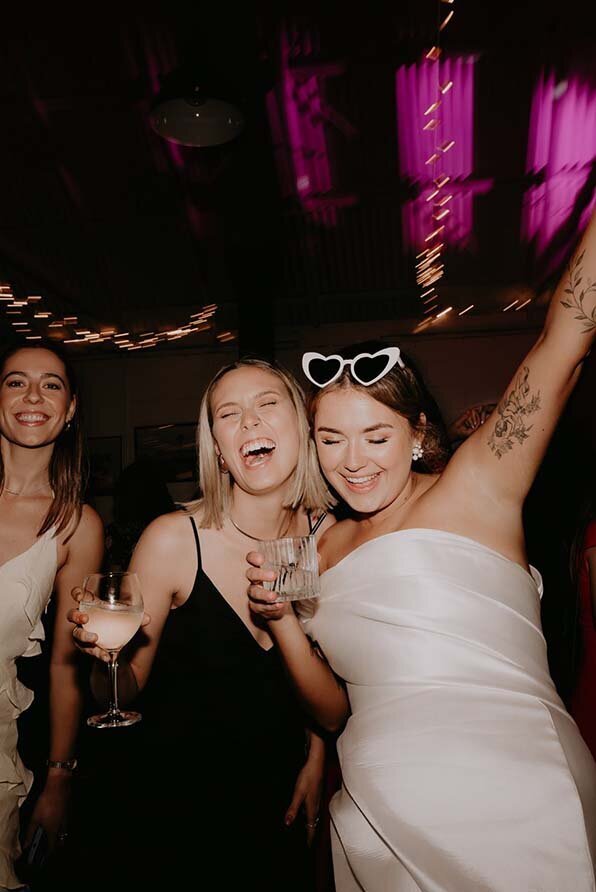 Bride in a white bridal dress and heart-shaped sunglasses celebrating with friends, holding drinks and surrounded by party lights.