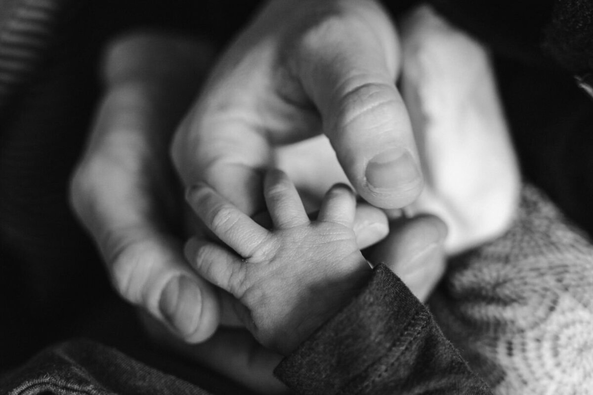 Black-and-white close-up of an adult gently holding a newborn’s tiny hand, highlighting the tender contrast between the large fingers and the baby’s delicate skin.