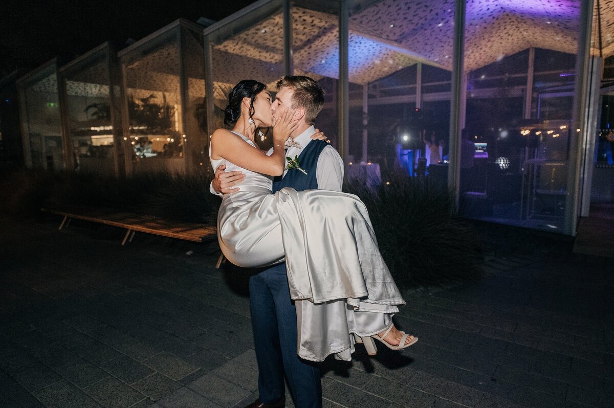 groom carrying bride at night outside ilex in christchurch botanic gardens purple lights in background