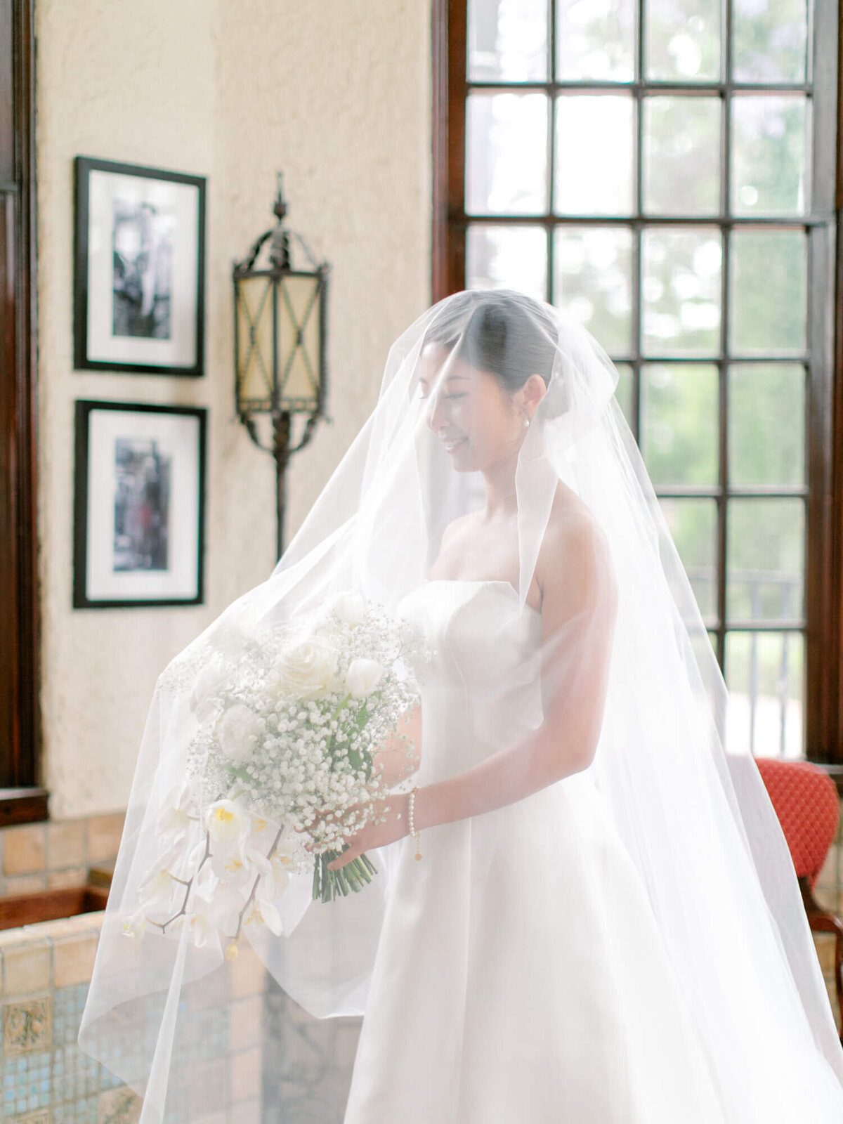 A bride in a flowing white gown and veil stands in a softly lit room, holding a bouquet of white flowers. The ambiance is elegant and serene, with vintage lanterns and framed photos on the walls.