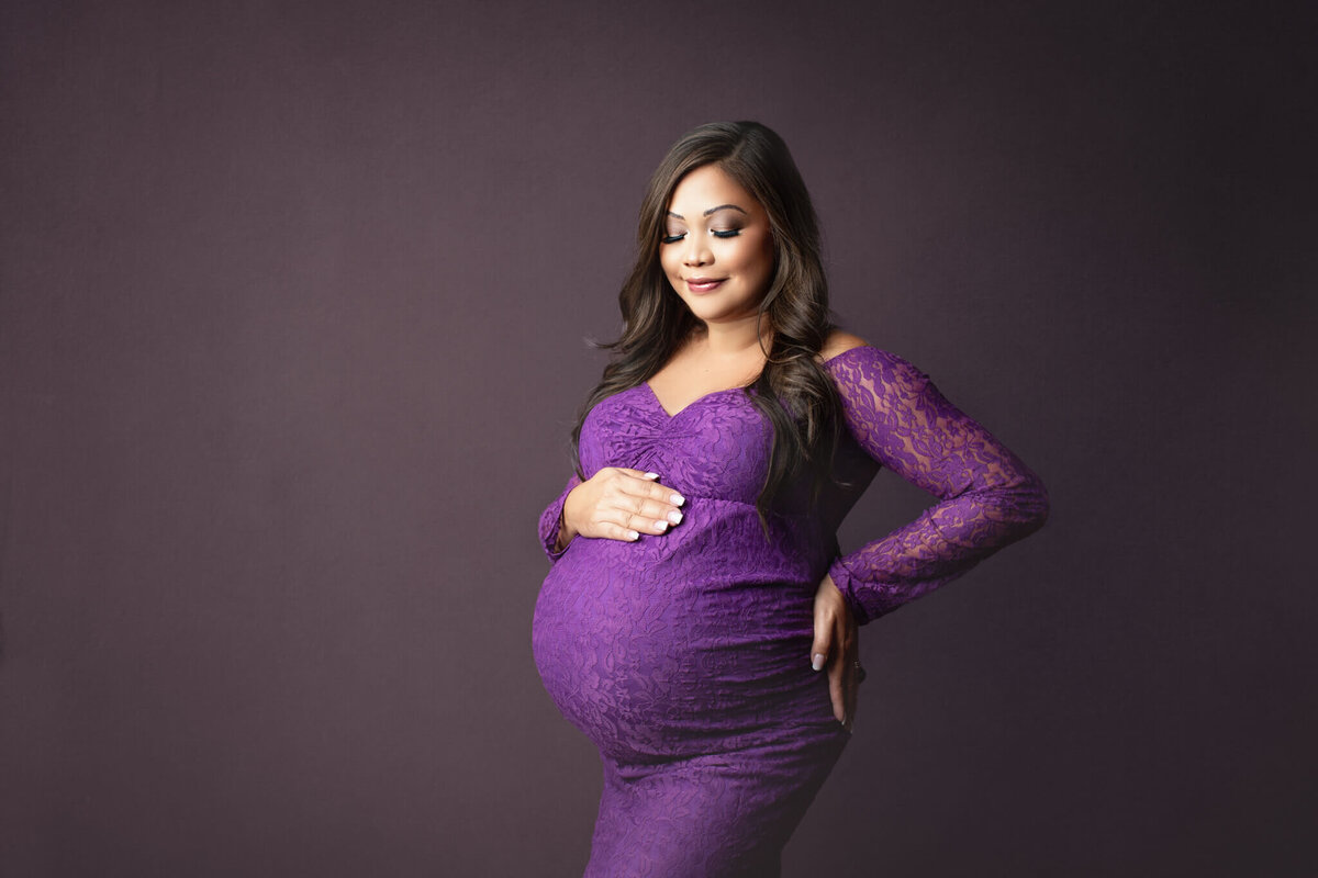 pregnant mom posed looking down at her belly in purple lace gown at niagara maternity studio