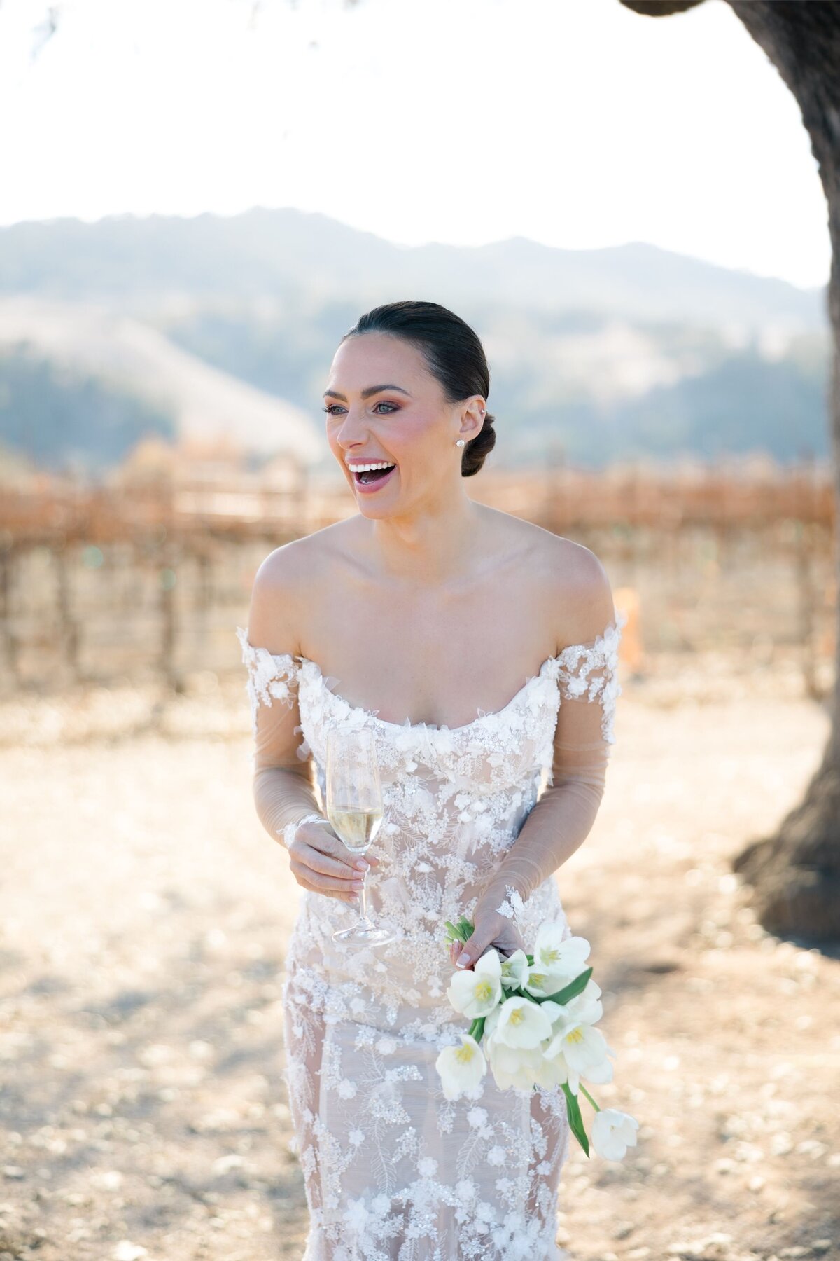 Bride smiling and laughing while holding a bouquet and champagne, surrounded by a vineyard landscape.
