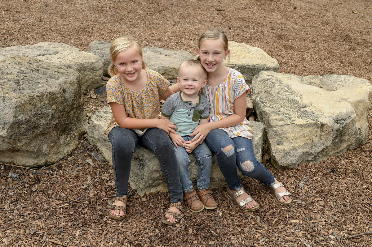 Three children sitting on rocks in an outdoor setting surrounded by trees and greenery