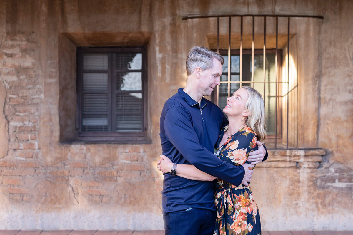 A husband with blonde hair wearing a navy long sleeve shirt hugging and looking down at his wife with blonde hair wearing an orange and navy floral dress looking up at him with her arms around his waist.